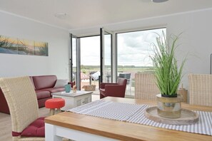 Ein modernes Wohnzimmer mit einer Couch, Sesseln, einem Esstisch und einem großen Fenster mit Blick ins Freie.