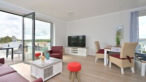 A modern living room with a sofa, red armchair, and a dining area with wicker chairs. A flat-screen TV is mounted on the wall. - Holiday apartment in Glowe with swimming pool (Glowe)
