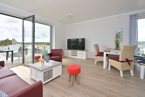 A modern living room with a sofa, red armchair, and a dining area with wicker chairs. A flat-screen TV is mounted on the wall. - Holiday apartment in Glowe with swimming pool (Glowe)