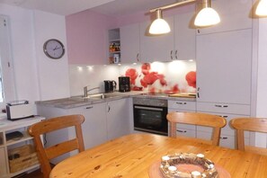 Eine kompakte Küche mit weißen Schränken, einem Holztisch und einer Uhr an der Wand.