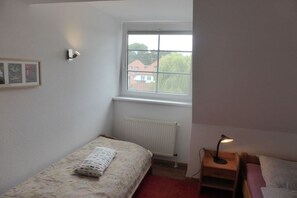 Ein kleines Schlafzimmer mit einem Einzelbett, einem Nachttisch mit Lampe, einem Fenster mit Blick auf Gebäude und einer Heizung.