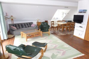 Ein Wohnzimmer mit einer Couch, Sesseln, einem Couchtisch, einem Esstisch mit Stühlen, einem Fernseher und einem Bücherregal.