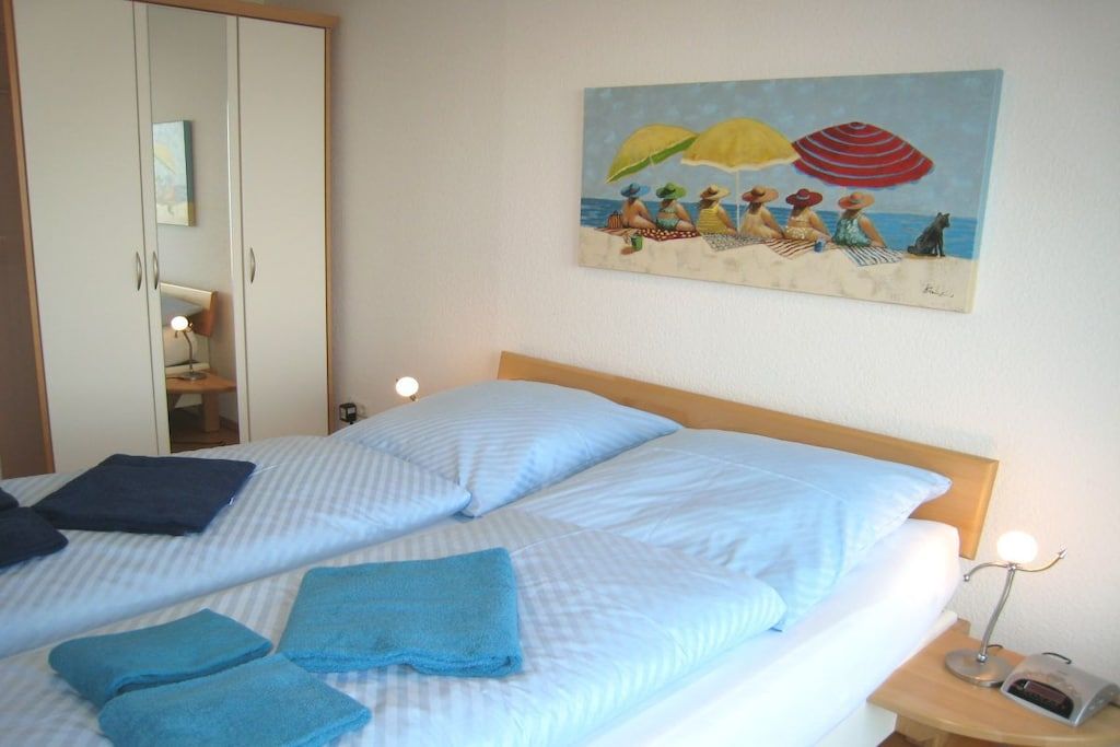 A hotel room with a bed, towels, a painting of a beach scene, a wardrobe, and a nightstand with a lamp.