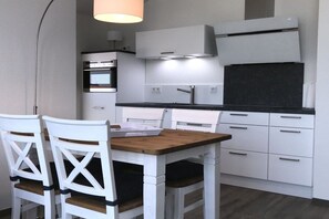 Eine kompakte Küche mit weißen Schränken, einem eingebauten Ofen und einer schwarzen Arbeitsplatte. Zwei weiße Stühle mit Holzsitzen und -lehnen sind vorhanden.