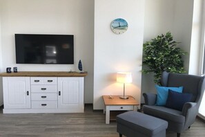 Ein modernes Wohnzimmer mit Fernseher, Holzschrank, Beistelltisch mit Lampe, grauem Sessel und einer Zimmerpflanze.