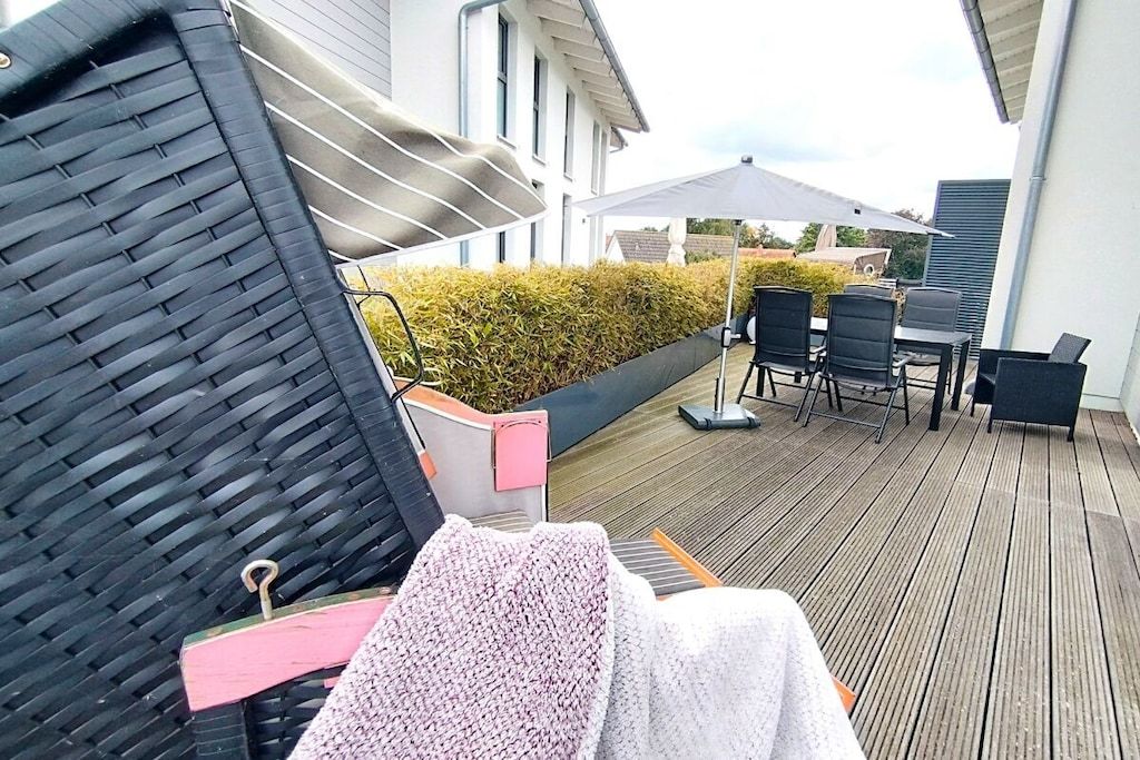 Ein überdachter Außenbereich mit Tisch und Stühlen, einem schwarzen Sonnenschirm und einer Hecke im Hintergrund.