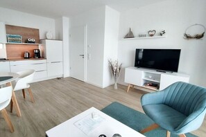 A modern living room with a flat-screen TV, a blue armchair, a white coffee table, and a dining area with a white table and chairs. - Holiday apartment in Glowe with swimming pool (Glowe)