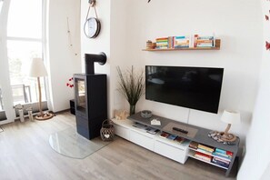 A modern living room with a flat-screen TV, a fireplace, a bookshelf, and a window with a view. - Holiday apartment in Glowe with swimming pool (Glowe)