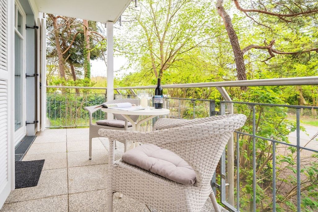 Ein Balkon mit Korbmöbeln, einem Tisch und einem Stuhl, mit Blick auf einen Garten mit Bäumen und einem Zaun.