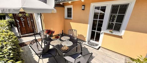 Eine Terrasse im Freien mit Tisch und Stühlen, einem weißen Sonnenschirm und einem Haus mit einem Ziegeldach.