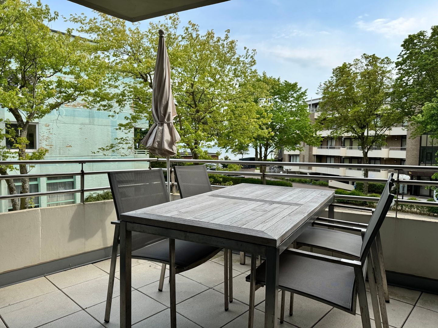 Eine Terrasse im Freien mit Tisch und Stühlen, einem beschatteten Sonnenschirm und Blick auf Gebäude und Bäume.