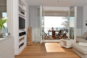 A modern living room with a wooden floor, a white sofa, a dining area with a table and chairs, and a view through a glass door to the outside. - Holiday apartment with 2 bedrooms (Sassnitz)