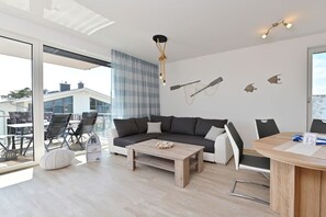 A modern living room with a sofa, coffee table, and chairs. There is a dining area with a table and chairs. The room has large windows and a view of the outside. - The sea is always a good idea! (Glowe)