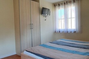 Ein Schlafzimmer mit einem Bett, einem Kleiderschrank, einem an der Wand montierten Fernseher und einem Fenster mit Vorhängen.