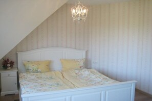 A bedroom with a sloped ceiling, a white bed with floral bedding, a nightstand with a vase of flowers, and a chandelier. - Country House Lake District 2 (Lychen)