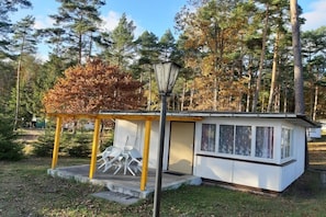 Een klein huisje met een veranda, een tafel en stoelen, omgeven door bomen.