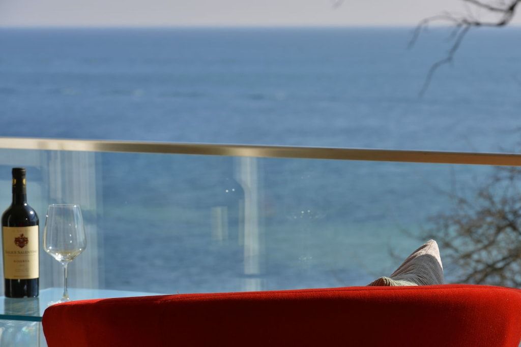 A glass table with a red chair, a bottle of wine, and a glass. A view of the ocean is visible through the glass.