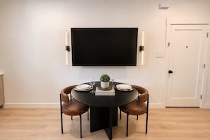 A dining area with a round table, four chairs, a flat-screen TV mounted above, and a door to the right.