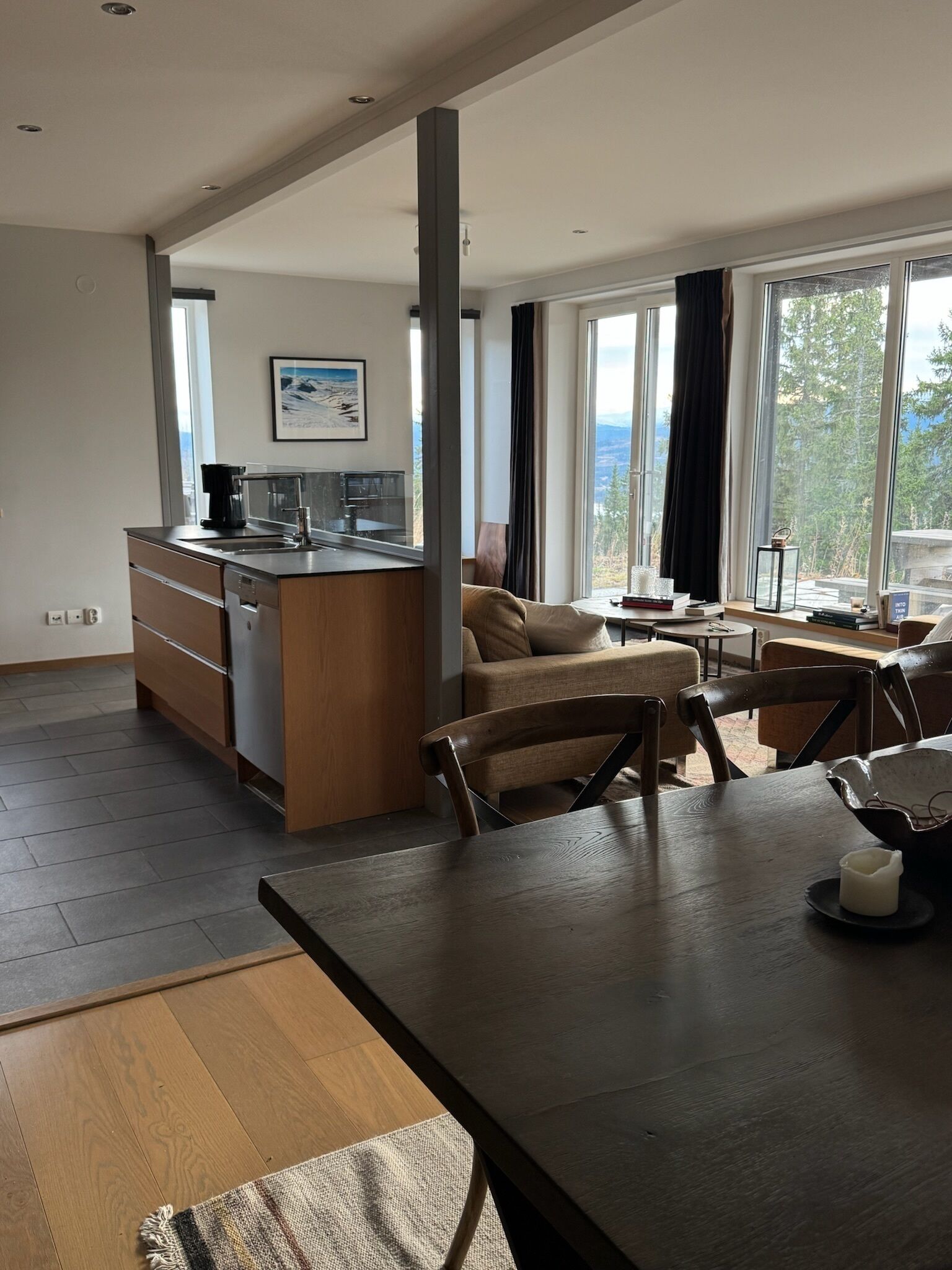 A modern kitchen with a central island, a dining area with a table and chairs, and large windows offering a view of the outdoors.