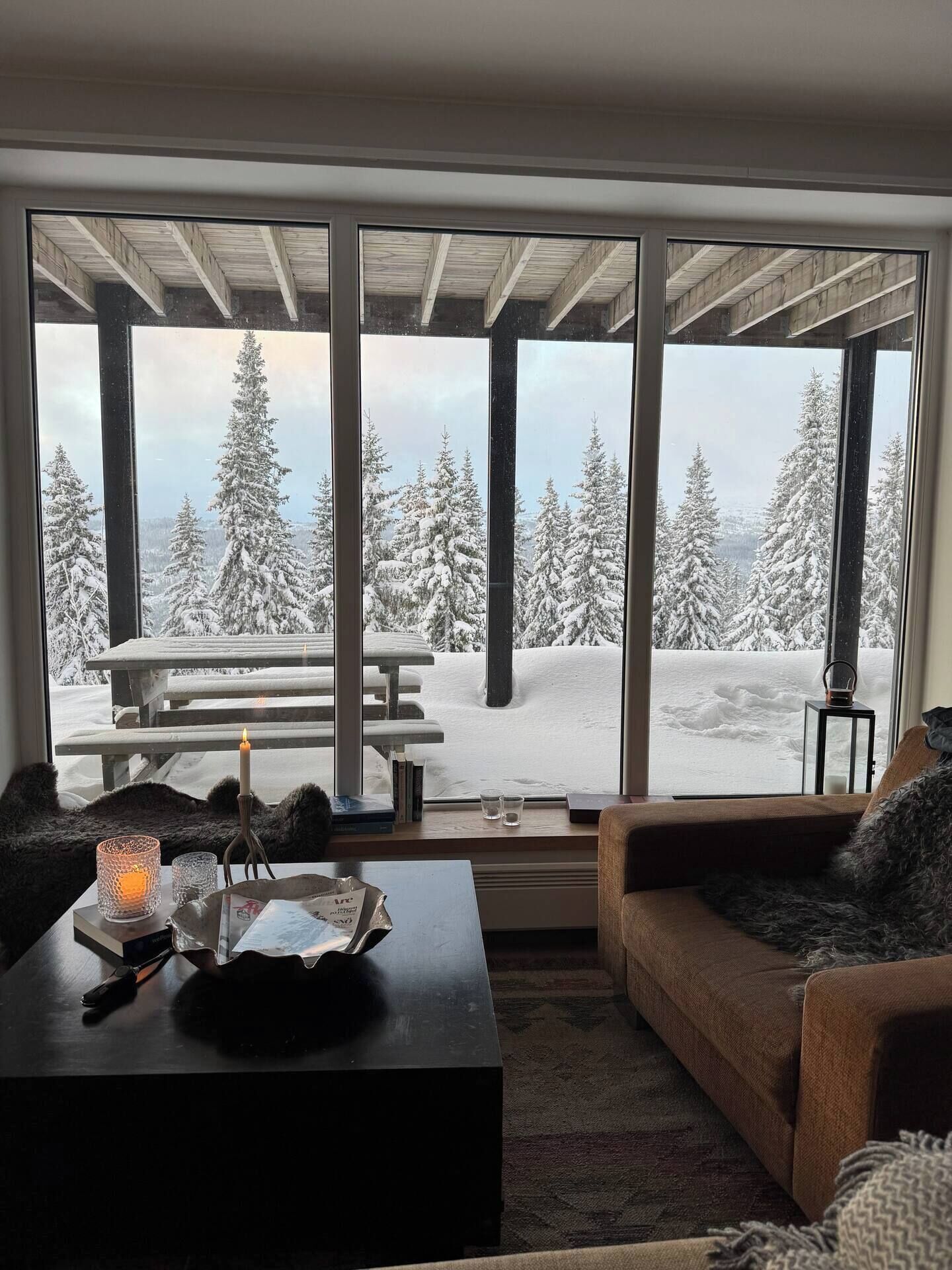 Ett mysigt vardagsrum med ett stort fönster med utsikt över ett snötäckt landskap, en träbänk utanför och en öppen spis med ett ljus.