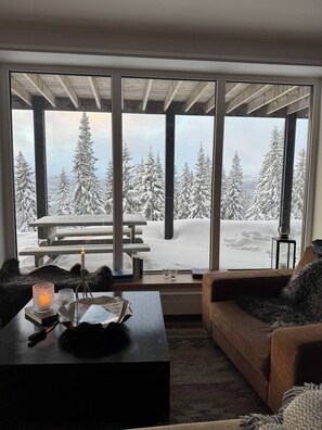 A cozy living room with a large window overlooking a snowy landscape, a wooden bench outside, and a fireplace with a candle.