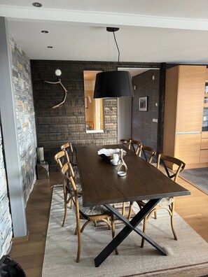 Ein Essbereich mit einem langen Holztisch, sechs Stühlen, einer schwarzen Pendelleuchte und einer Steinwand.