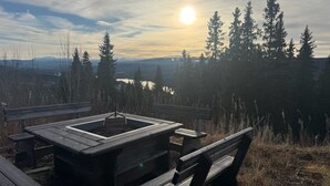 Een houten picknicktafel met banken, een vuurkuil en een uitzicht op een meer en beboste bergen op de achtergrond.