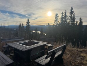 Ein Holztisch mit Bänken, eine Feuerstelle und ein Blick auf einen See und bewaldete Berge im Hintergrund.