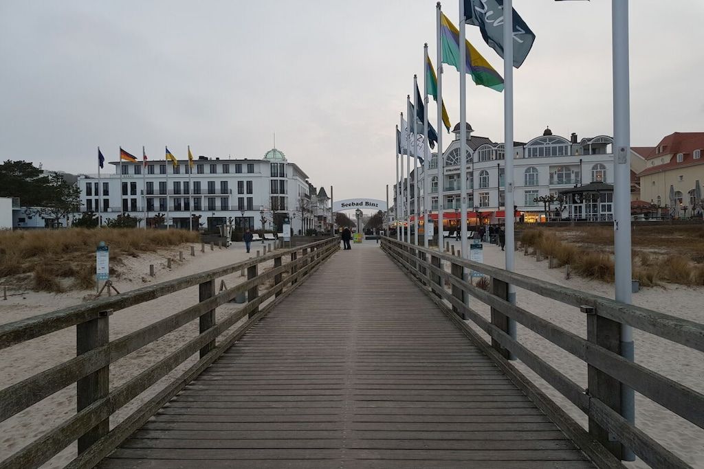 A wooden boardwalk leading to a building with multiple flags, a sandy beach, and a cloudy sky.