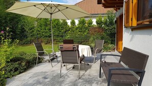 En uteplats med ett bord och stolar, ett stort parasoll och en byggnad med ett tak täckt av tegelpannor i bakgrunden.