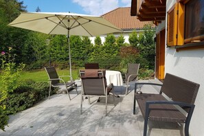 Een terras buiten met een tafel en stoelen, een grote parasol en een gebouw met een pannendak op de achtergrond.