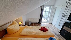 Una camera d'albergo con soffitto spiovente, un letto con biancheria gialla, un panno rosso sul letto, una lampada su un comodino, un termosifone e un mobile TV.