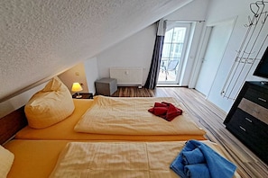 Quarto de hotel com teto inclinado, cama com roupa de cama amarela, um tecido vermelho na cama, abajur em uma mesa de cabeceira, radiador e um suporte para TV.