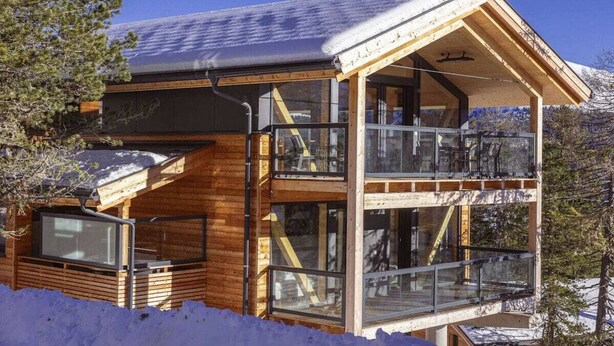 Uma cabana de madeira com varanda e janelas amplas, rodeada por neve e árvores.