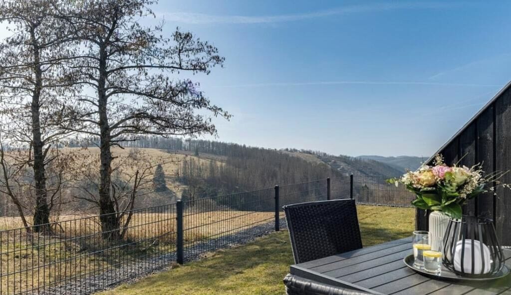 A table with a vase of flowers and a glass of water, a chair, and a view of a fenced area with trees and hills.