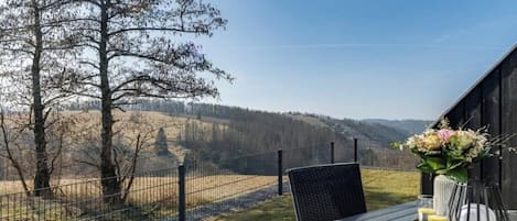 A table with a vase of flowers and a glass of water, a chair, and a view of a fenced area with trees and hills.