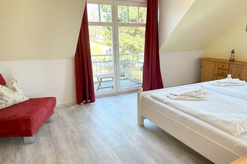 Ein Schlafzimmer mit einem Bett, einem roten Schlafsofa, einem Holzkasten und einem Balkon mit Tisch und Stühlen.