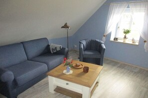 Ein Zimmer mit einem blauen Sofa, einem Holztisch, einer Stehlampe und einem Fenster mit Vorhängen.