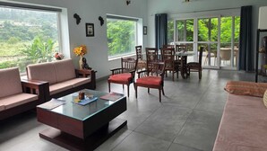 A spacious living room with a glass coffee table, sofas, and a dining area with chairs. Large windows offer a view of greenery outside.