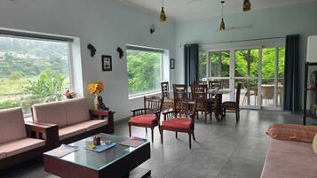 A spacious living room with a glass coffee table, sofas, and a dining area with chairs. Large windows offer a view of greenery outside.