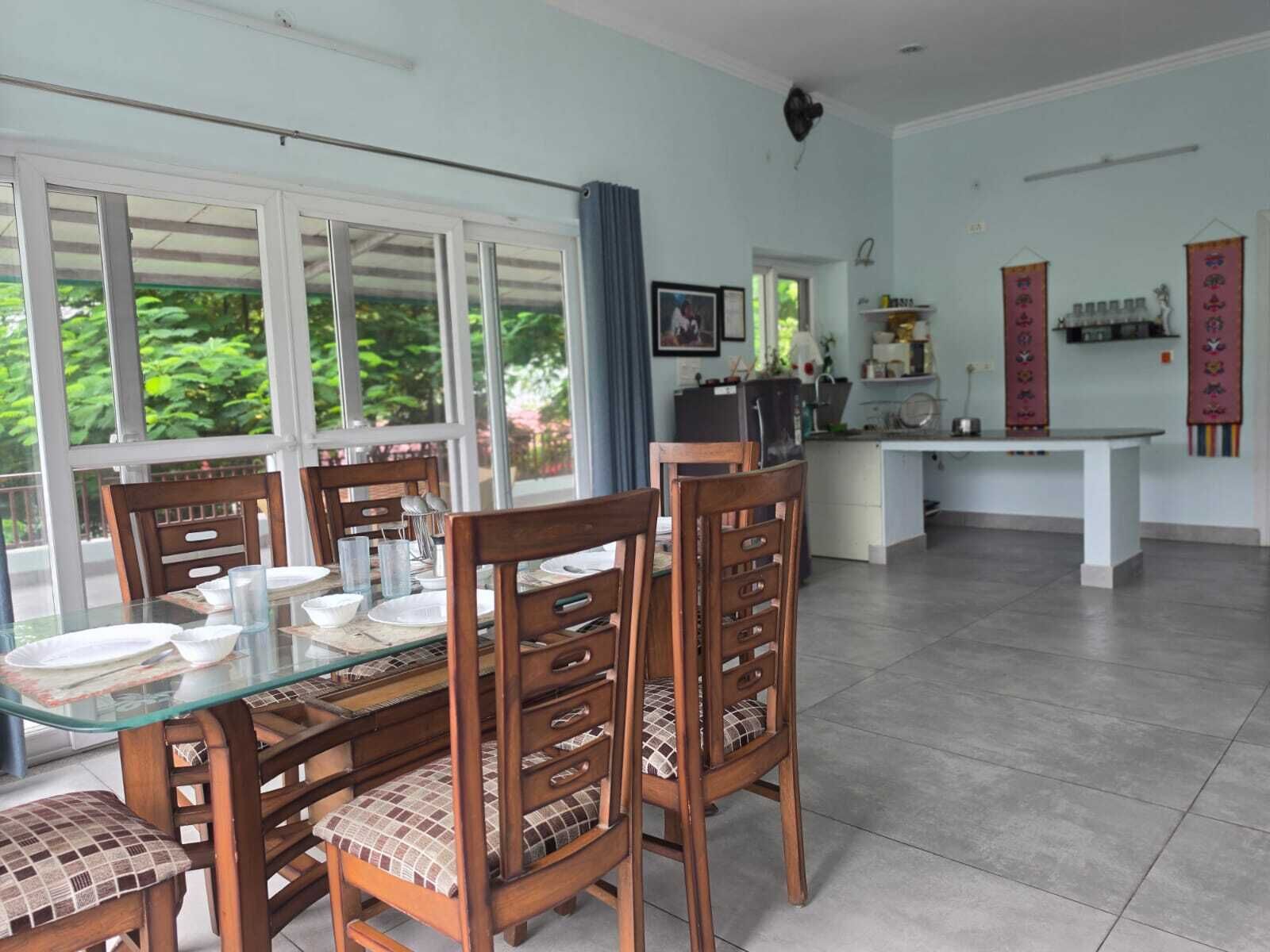 Un comedor con mesa de vidrio y sillas de madera, una cocina al fondo y ventanas grandes con vistas a zonas verdes.