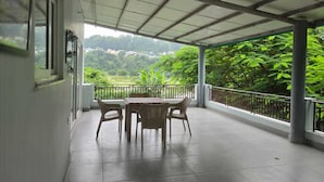 Uma área externa coberta com mesa, cadeiras, corrimão e vista para uma paisagem verde.