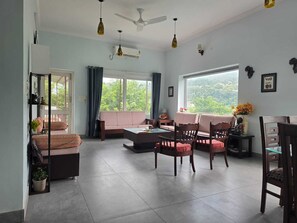 A spacious living area with a sofa, chairs, a coffee table, and a dining table. There are ceiling fans, a large window with a view, and a wall-mounted air conditioner.