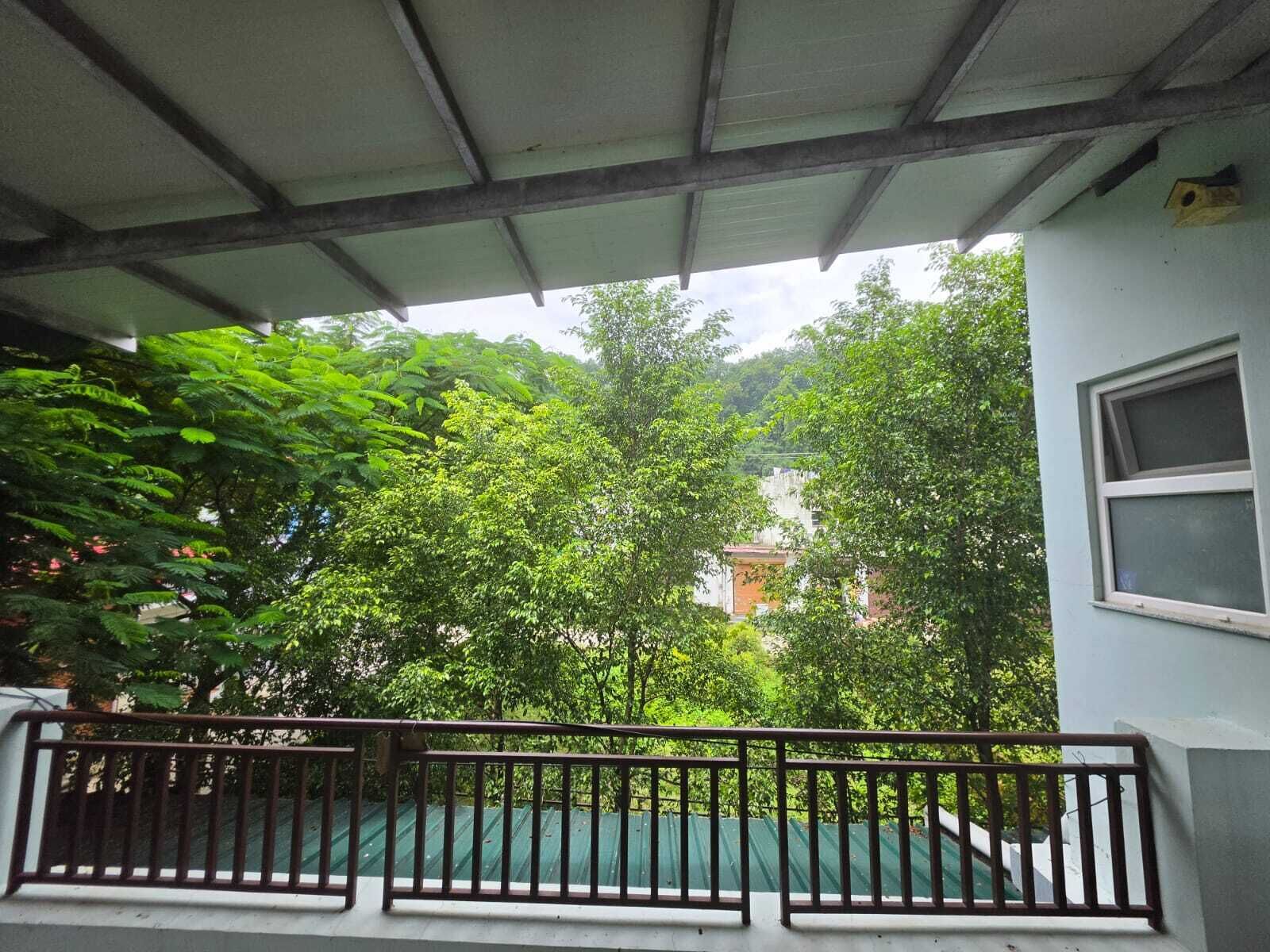 A covered balcony with a metal railing, overlooking a lush green area with trees and a building in the background.
