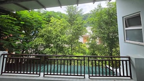 A covered balcony with a metal railing, overlooking a lush green area with trees and a building in the background.