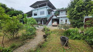 Uma casa de dois andares com um caminho de cascalho que leva à entrada, cercada por vegetação exuberante.