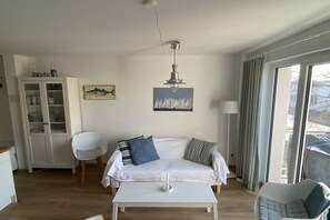 A living room with a white sofa, a white coffee table, a cabinet with glass doors, and a painting of sailboats. - Baltic Sea Oasis Rerik (Ostseebad Rerik)