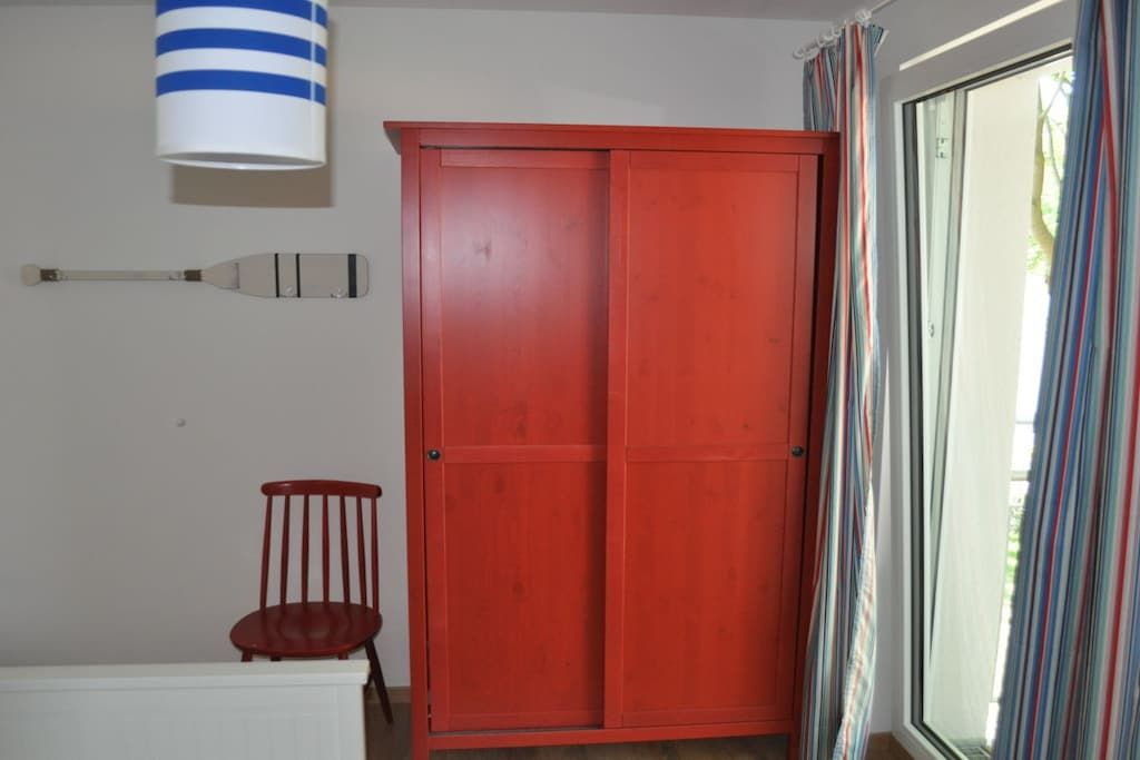 A room with a red wardrobe, a white chair, a striped lamp, and a window with curtains.