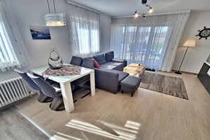 Ein modernes Wohnzimmer mit einer Sofagarnitur, einem Esstisch mit Stühlen, einer Lampe und Blick ins Freie durch große Fenster.