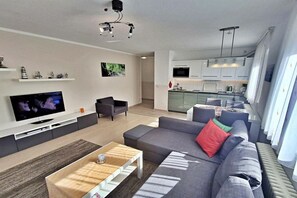 Ein modernes Wohnzimmer mit Fernseher, grauem Sofa, Couchtisch und einer Küche im Hintergrund.